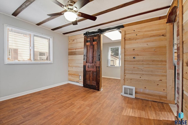 unfurnished bedroom with ceiling fan, a barn door, beamed ceiling, light hardwood / wood-style floors, and wooden walls