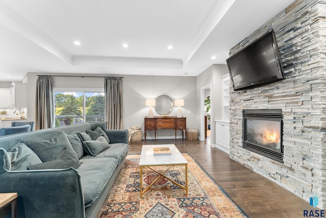 living room featuring a tray ceiling, a stone fireplace, crown molding, and dark wood-type flooring