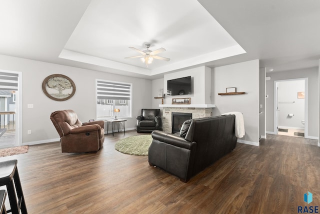 living room with ceiling fan, dark hardwood / wood-style floors, a stone fireplace, and a raised ceiling