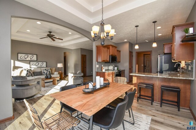 dining area featuring light wood-type flooring, ceiling fan with notable chandelier, a tray ceiling, and sink