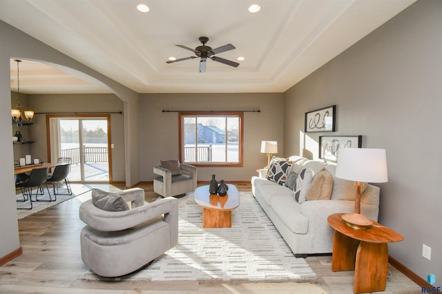 living room with plenty of natural light, light wood-type flooring, ceiling fan with notable chandelier, and a tray ceiling