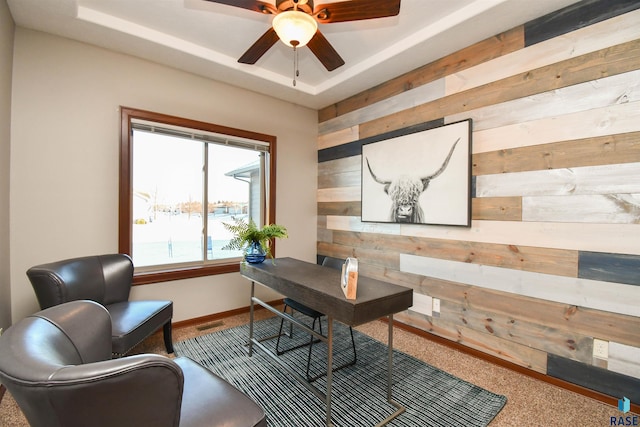 carpeted office space with ceiling fan, wood walls, and a raised ceiling