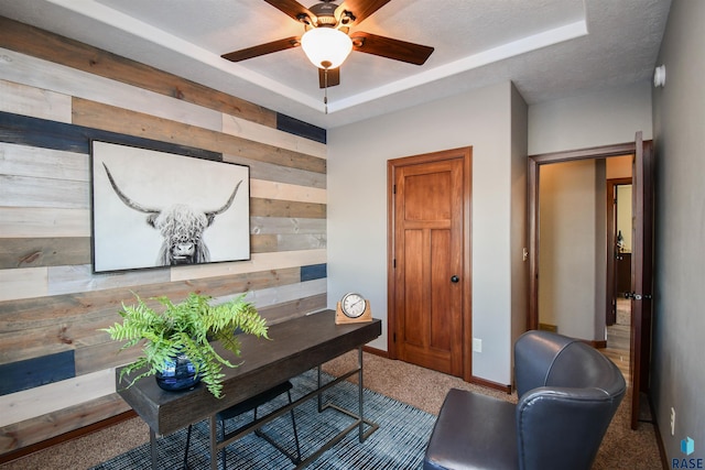 carpeted office space with a raised ceiling, wood walls, ceiling fan, and a textured ceiling