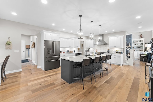 kitchen with high quality fridge, white cabinets, wall chimney exhaust hood, and an island with sink