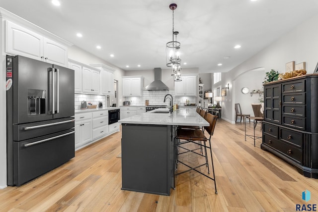 kitchen with black appliances, an island with sink, wall chimney exhaust hood, sink, and white cabinetry