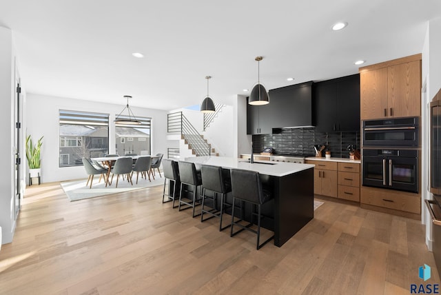 kitchen featuring decorative light fixtures, an island with sink, wall chimney range hood, and light hardwood / wood-style flooring