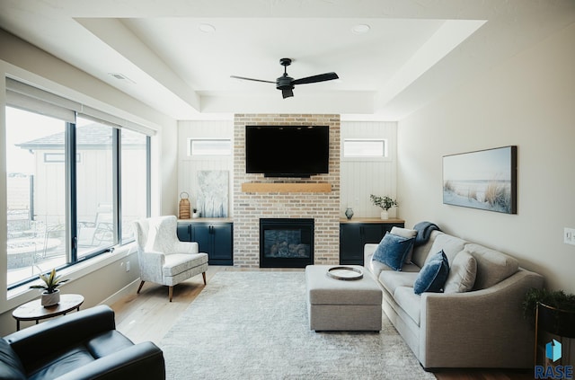 living room with a fireplace, a raised ceiling, ceiling fan, and light wood-type flooring