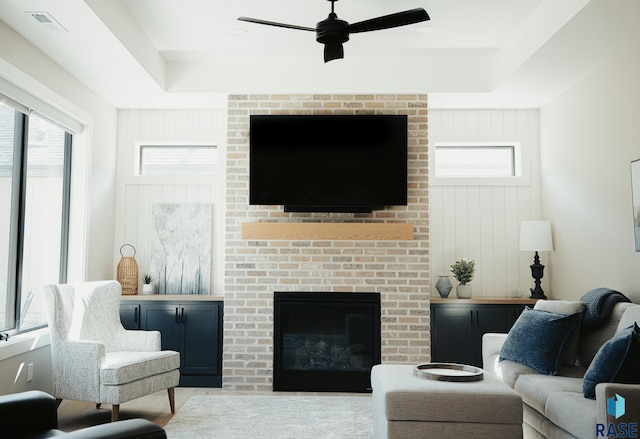 living room with ceiling fan, a brick fireplace, and a tray ceiling