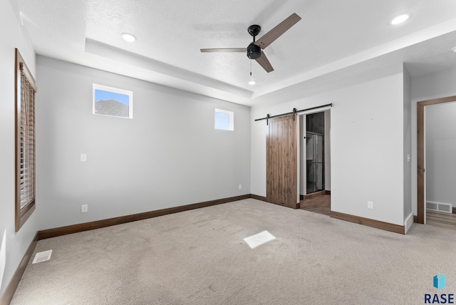 unfurnished bedroom featuring a barn door, ceiling fan, a tray ceiling, light colored carpet, and a textured ceiling