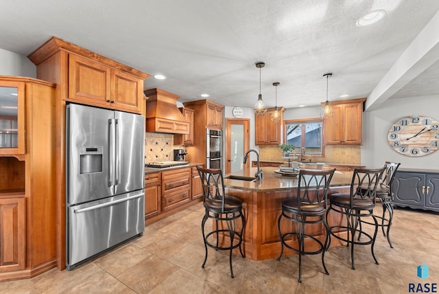 kitchen featuring appliances with stainless steel finishes, dark stone countertops, an island with sink, sink, and premium range hood