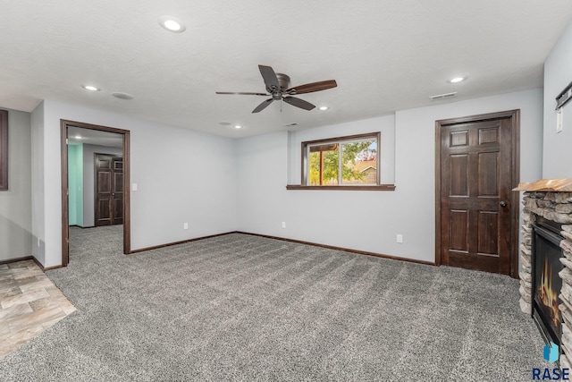 unfurnished living room with ceiling fan, a stone fireplace, a textured ceiling, and carpet floors