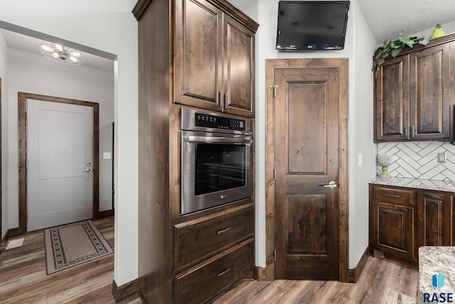 kitchen with dark brown cabinetry, wood-type flooring, light stone countertops, decorative backsplash, and stainless steel oven