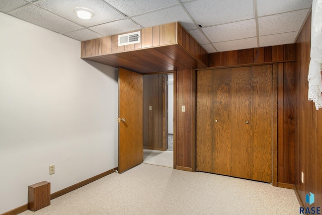 unfurnished bedroom with light carpet, a drop ceiling, wooden walls, and a closet