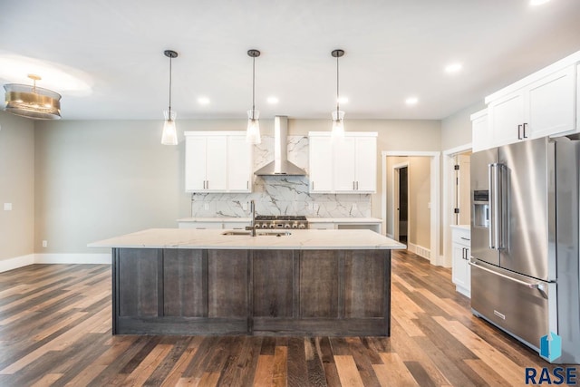 kitchen with wall chimney range hood, sink, white cabinetry, high quality fridge, and an island with sink