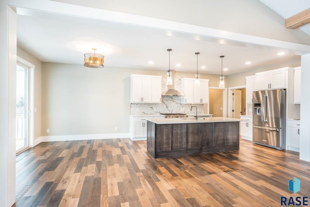 kitchen featuring high end fridge, a center island with sink, hanging light fixtures, wall chimney range hood, and white cabinets