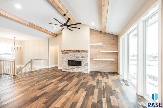 unfurnished living room with a stone fireplace, dark hardwood / wood-style floors, ceiling fan with notable chandelier, wooden walls, and vaulted ceiling with beams