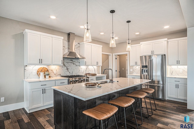 kitchen featuring wall chimney range hood, high end appliances, white cabinets, a center island with sink, and decorative light fixtures