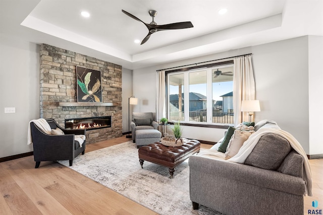 living room featuring ceiling fan, a tray ceiling, light hardwood / wood-style floors, and a stone fireplace