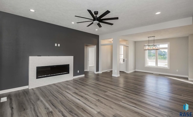 unfurnished living room featuring dark wood-type flooring, ceiling fan with notable chandelier, a brick fireplace, and a textured ceiling