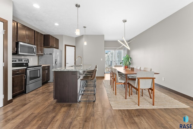 kitchen with appliances with stainless steel finishes, hanging light fixtures, dark brown cabinets, a center island with sink, and a kitchen bar