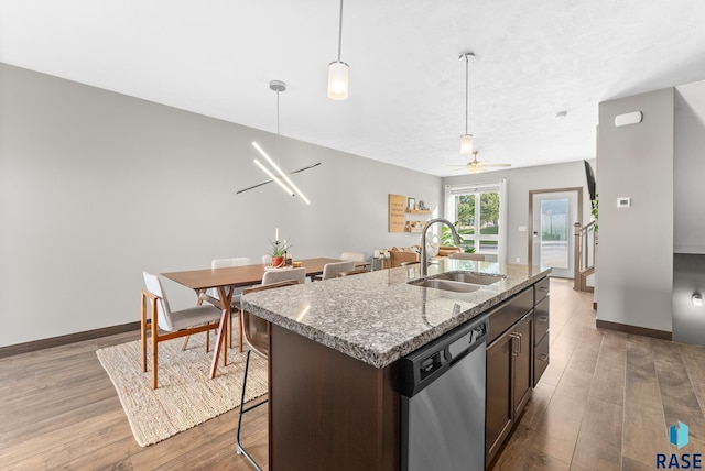 kitchen with dark brown cabinetry, sink, stainless steel dishwasher, an island with sink, and light stone countertops