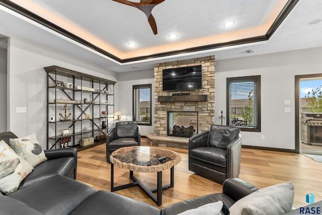 living room featuring ceiling fan, a tray ceiling, a stone fireplace, and light wood-type flooring