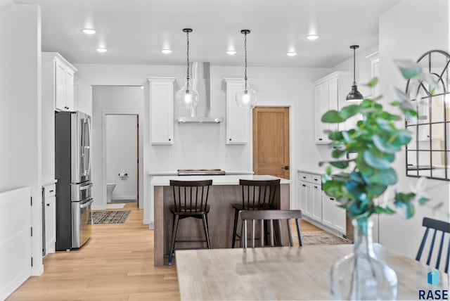 kitchen with white cabinetry, wall chimney range hood, stainless steel fridge, and hanging light fixtures