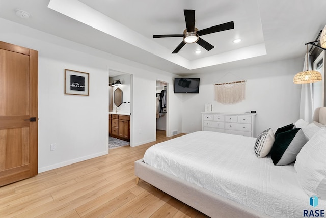 bedroom with ceiling fan, connected bathroom, a tray ceiling, a walk in closet, and light wood-type flooring