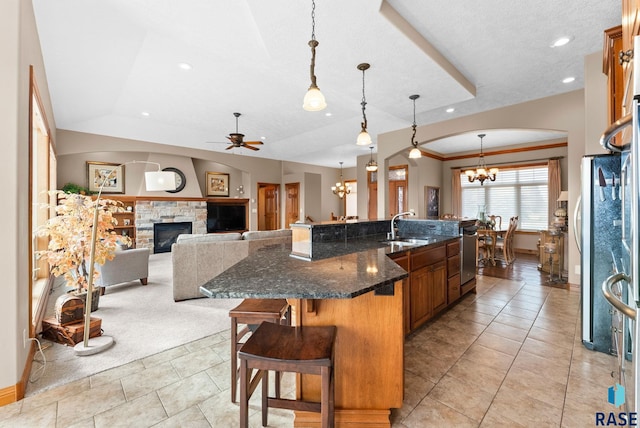 kitchen featuring a kitchen bar, a stone fireplace, decorative light fixtures, dark stone countertops, and a large island with sink