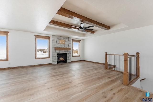 unfurnished living room featuring a stone fireplace, a tray ceiling, ceiling fan, light hardwood / wood-style floors, and beam ceiling