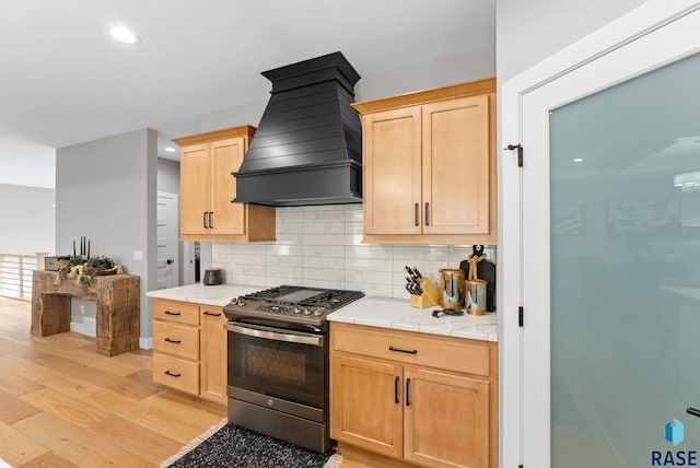 kitchen with light brown cabinetry, custom exhaust hood, stainless steel gas range oven, tasteful backsplash, and light wood-type flooring