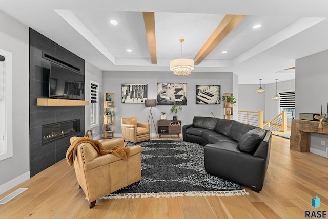 living room featuring a tiled fireplace, beam ceiling, an inviting chandelier, and light wood-type flooring