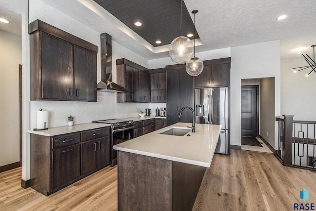 kitchen featuring sink, a raised ceiling, an island with sink, stainless steel appliances, and wall chimney range hood