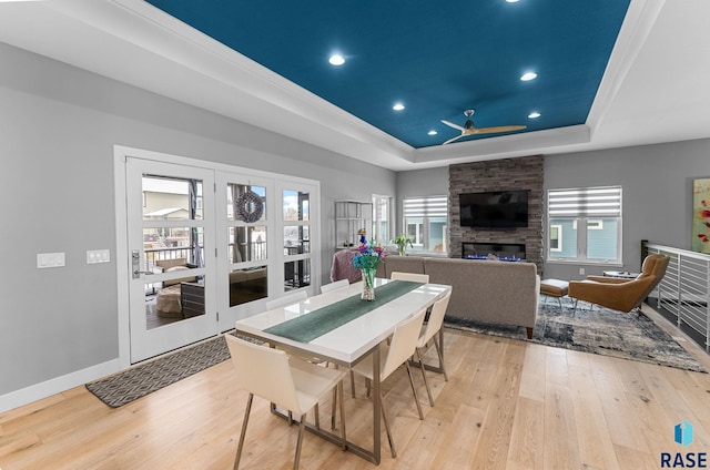 dining room with a stone fireplace, a raised ceiling, ceiling fan, and light wood-type flooring