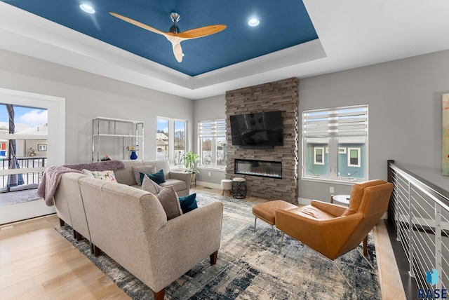 living room with a tray ceiling, a stone fireplace, light hardwood / wood-style floors, and ceiling fan