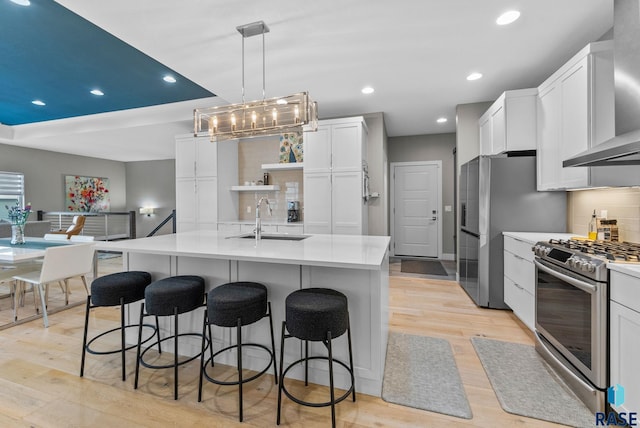 kitchen featuring wall chimney exhaust hood, white cabinetry, decorative light fixtures, appliances with stainless steel finishes, and a kitchen island with sink