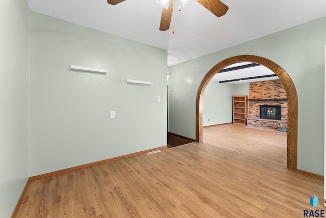 empty room featuring a brick fireplace, light hardwood / wood-style floors, beamed ceiling, and ceiling fan