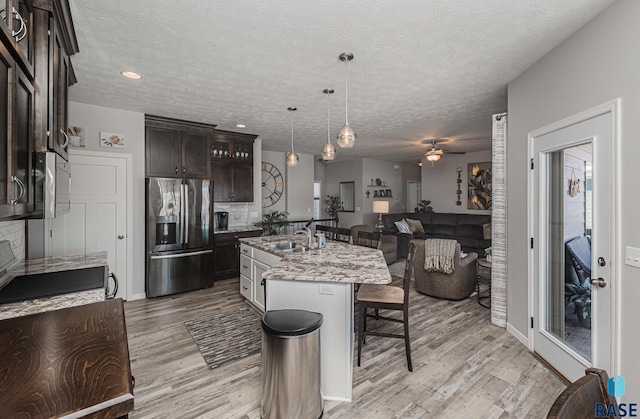 kitchen featuring a kitchen island with sink, hanging light fixtures, stainless steel refrigerator with ice dispenser, dark brown cabinets, and light stone counters