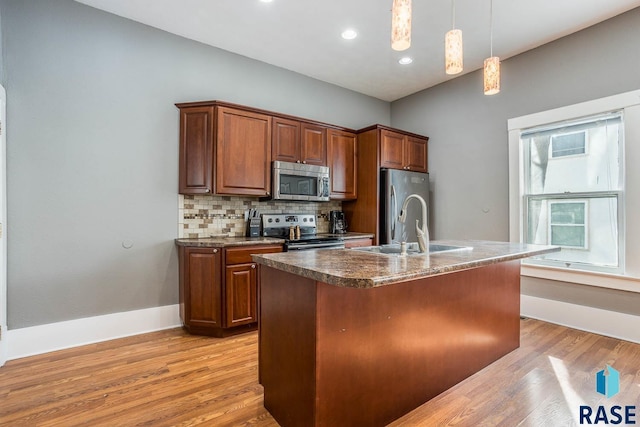 kitchen with dark countertops, appliances with stainless steel finishes, a center island with sink, a sink, and pendant lighting