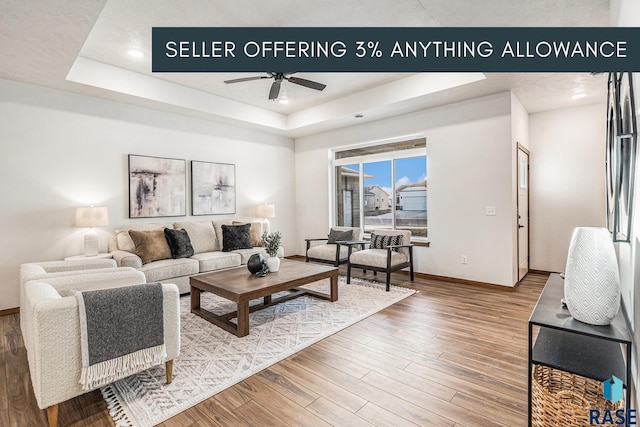 living area with ceiling fan, a tray ceiling, wood finished floors, and baseboards