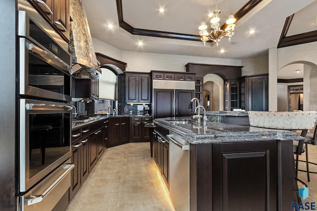 kitchen with a kitchen island with sink, appliances with stainless steel finishes, a raised ceiling, and arched walkways