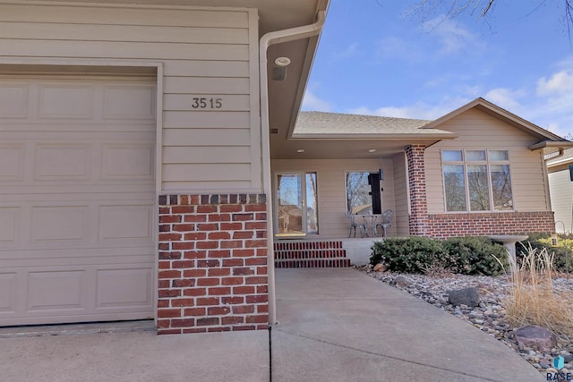 view of exterior entry featuring a shingled roof, brick siding, and an attached garage