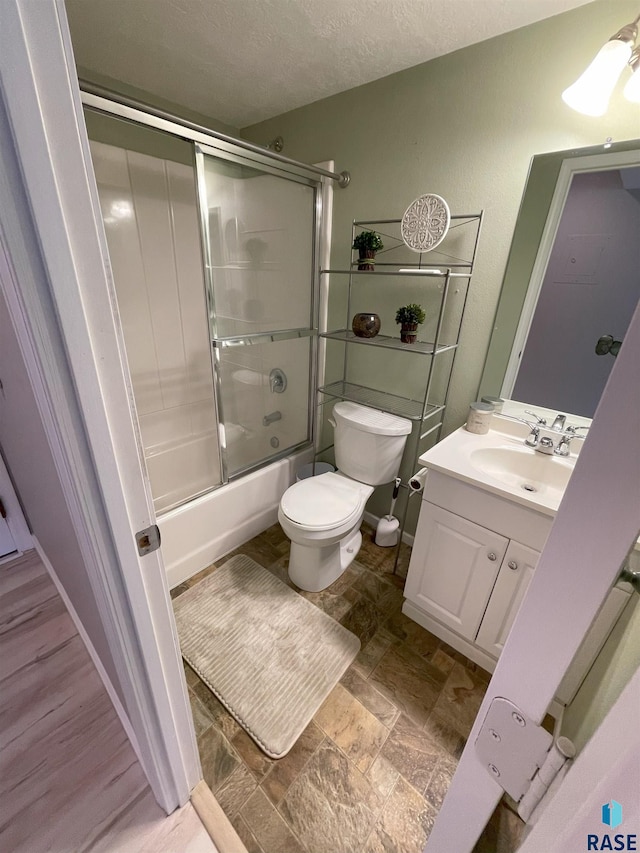 full bath featuring bath / shower combo with glass door, toilet, stone finish floor, a textured ceiling, and vanity