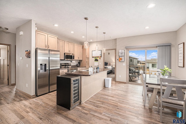 kitchen featuring dark countertops, appliances with stainless steel finishes, decorative light fixtures, a kitchen island with sink, and light brown cabinetry