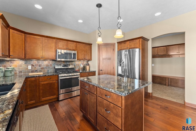 kitchen featuring dark wood finished floors, appliances with stainless steel finishes, brown cabinetry, a kitchen island, and dark stone countertops