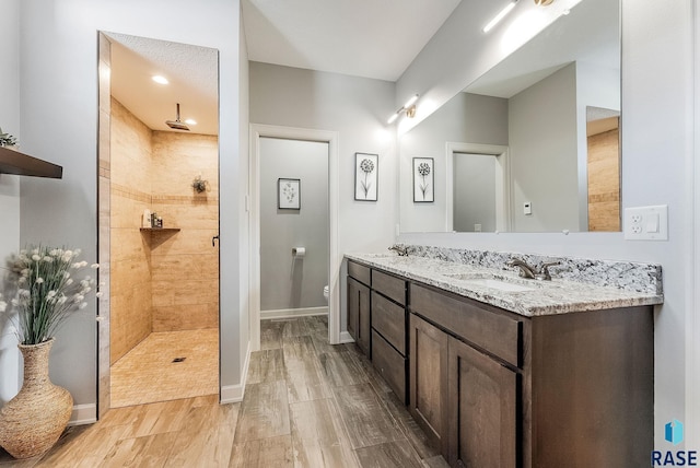 full bath with double vanity, a sink, a tile shower, wood finished floors, and baseboards