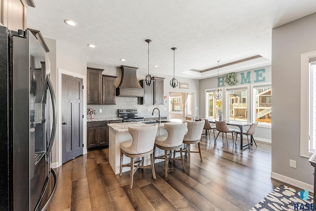 kitchen featuring a center island with sink, custom exhaust hood, stainless steel appliances, a raised ceiling, and hanging light fixtures