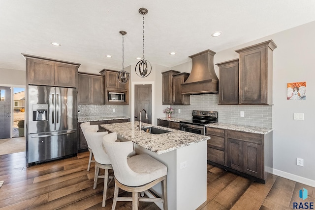 kitchen with appliances with stainless steel finishes, light stone counters, a kitchen island with sink, custom exhaust hood, and a sink