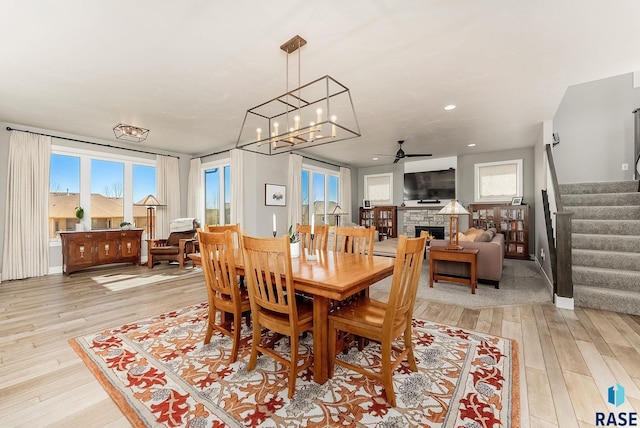 dining area featuring light wood finished floors, stairway, ceiling fan with notable chandelier, a stone fireplace, and recessed lighting