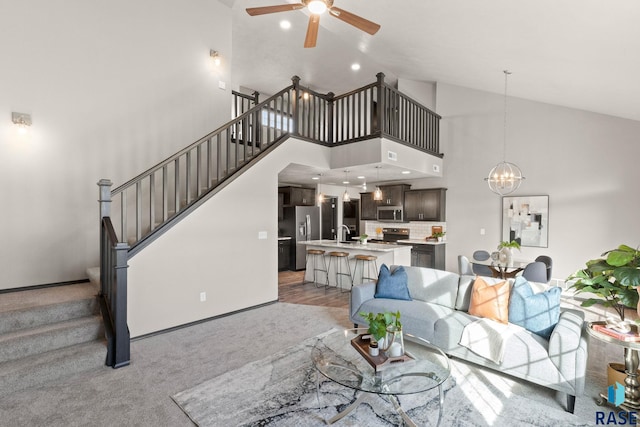 living area featuring light carpet, baseboards, stairway, high vaulted ceiling, and ceiling fan with notable chandelier